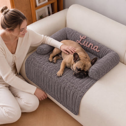A woman kneels beside a cream sofa, petting a sleeping fawn dog resting on a gray ribbed pet bed with a pink embroidered name that reads 'Luna'.